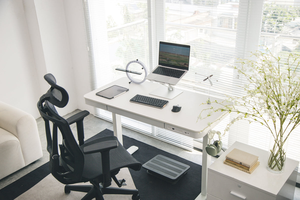 White minimalist standing desk by the window with a laptop on a stand, ring desk clock and black mesh chair