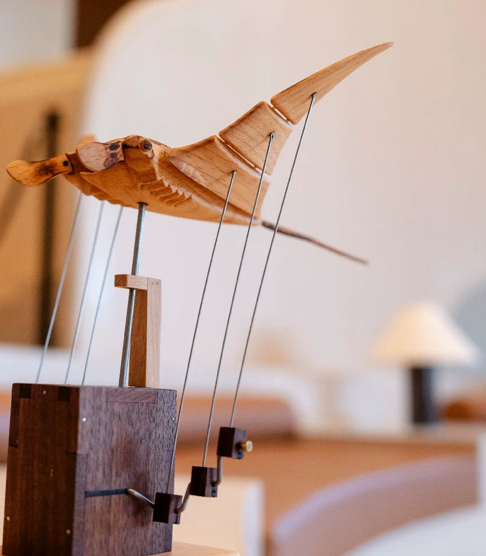 Light wood manta ray rising on fine wires above a dark wood box, a soft lamp and sofa blurred behind.