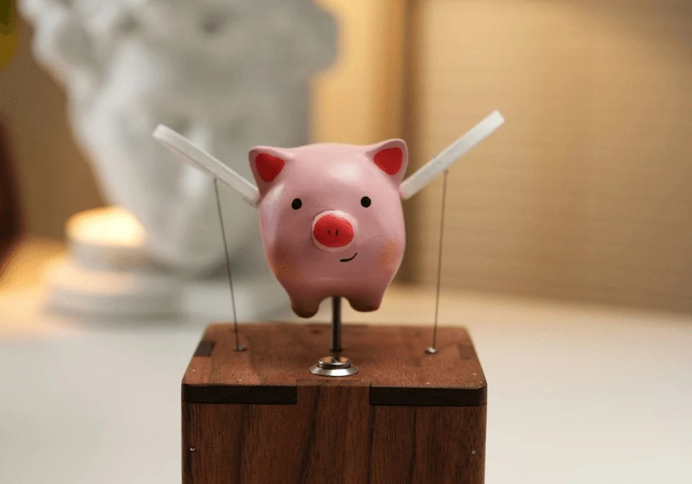 Front view of the pink pig with two white wings angled up on thin wires over the top of a reddish-brown wood box, a blurred plaster bust and a warm wall behind.