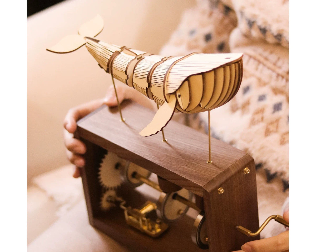 Two hands hold a dark wood-framed music box and turn its brass crank, a pale jointed wooden whale on brass rods above, a soft knit blanket behind.