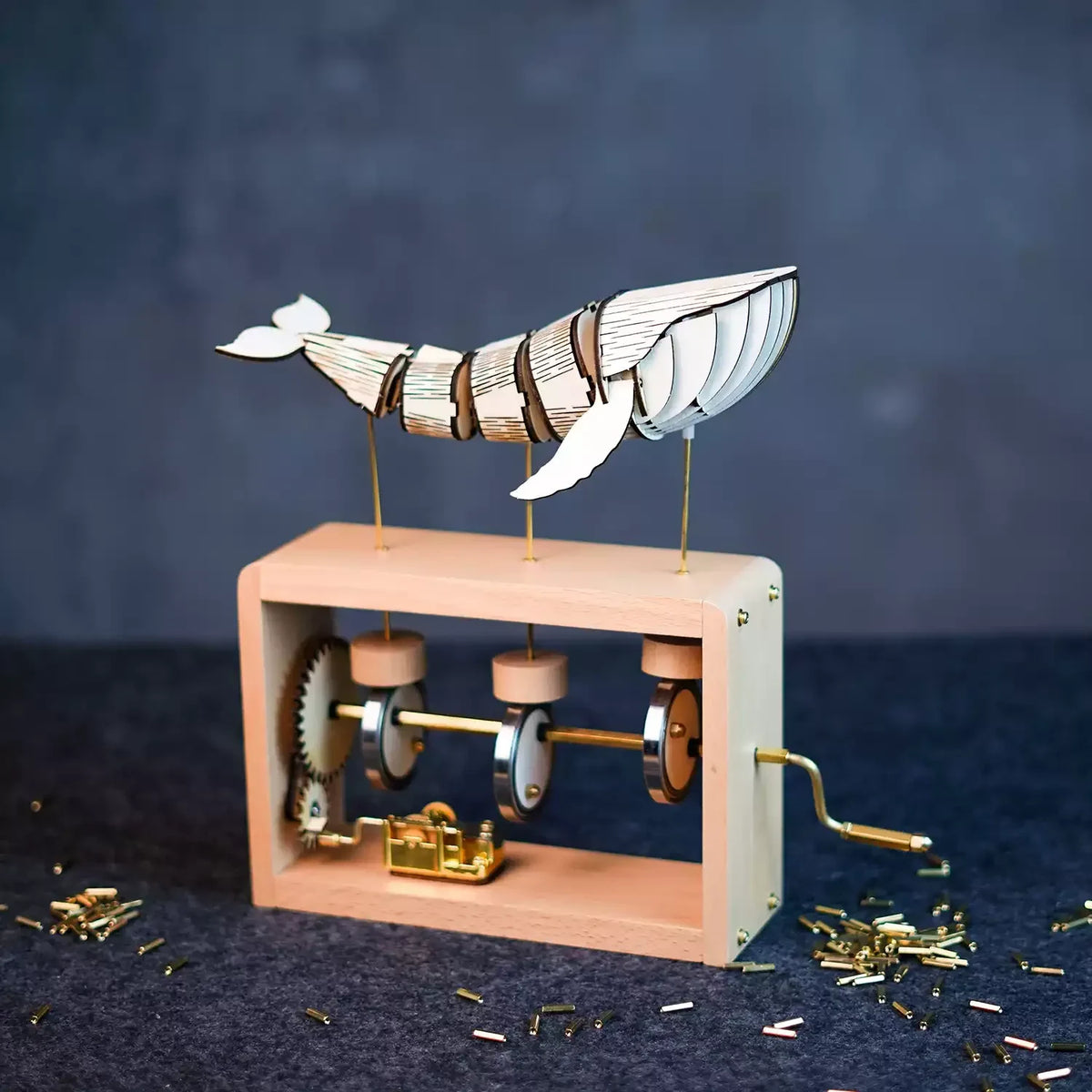 Pale wood edition on a dark blue cloth: a pale jointed wooden whale on three brass rods above a light wood frame with gears, cams and a gold music box.