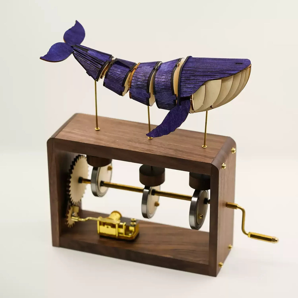 Dark wood edition on white: a blue-violet jointed wooden whale on three brass rods above a dark wood frame with gears, cams and a gold music box.
