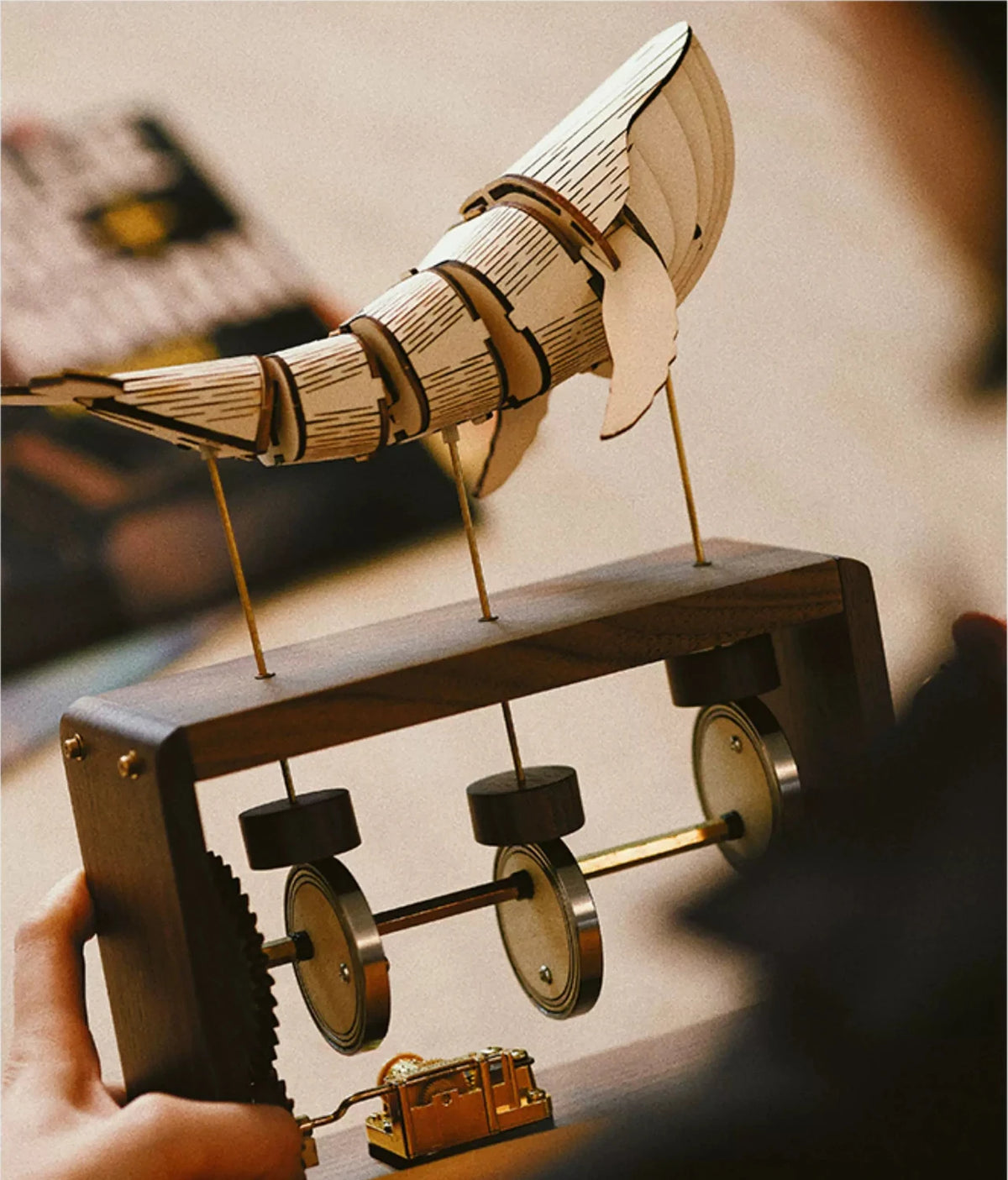 Hands hold a dark wood music box with a pale jointed whale on brass rods, three cams and the music box movement in view.