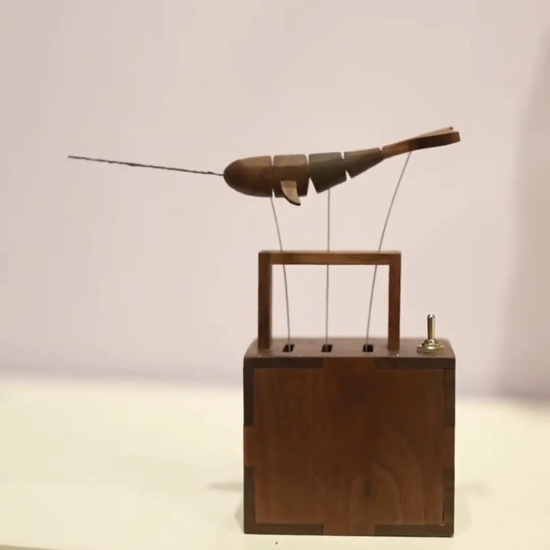 Front view of the dark wood whale hanging from three wires above an open dark wood frame on a finger-jointed dark wood box, small toggle switch at the right of the lid, pale wall behind.