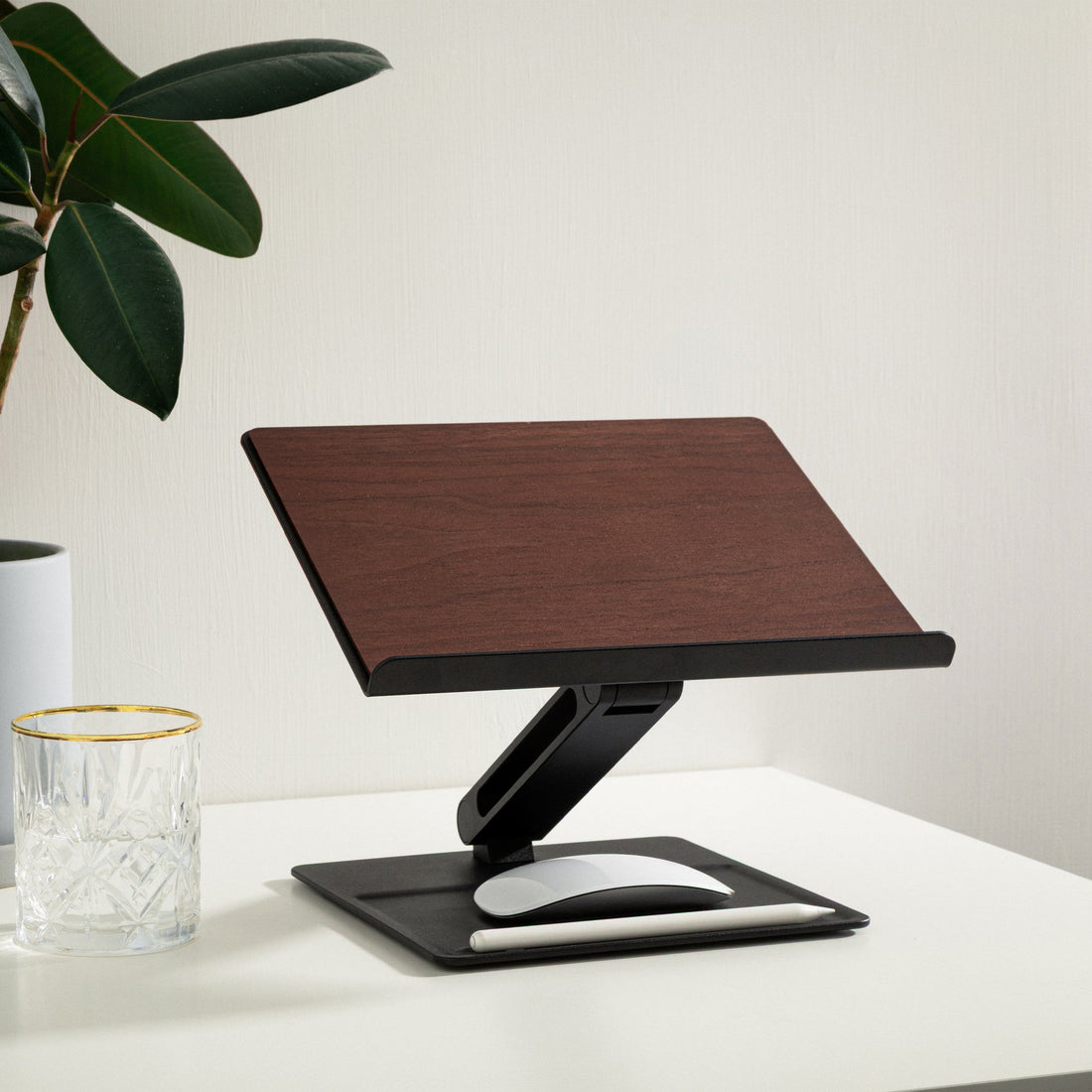Black and walnut stand on a white desk with a plant and a glass nearby, mouse resting on the base tray