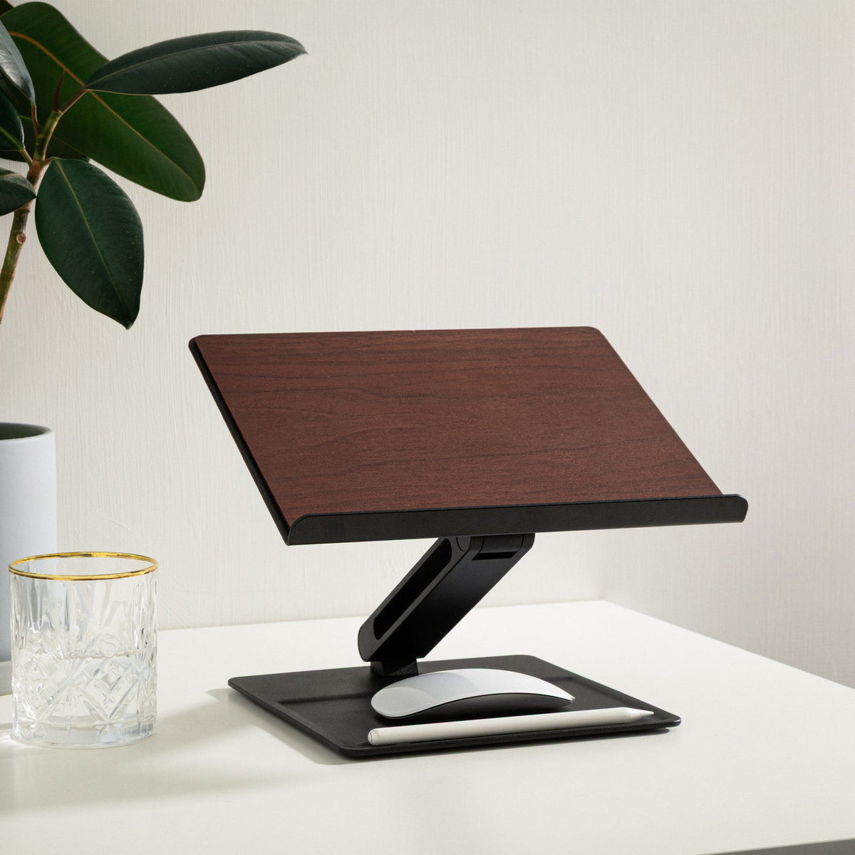 Black and walnut stand on a white desk with a plant and a glass nearby, mouse resting on the base tray