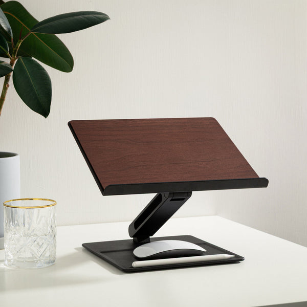 Black and walnut stand on a white desk with a plant and a glass nearby, mouse resting on the base tray