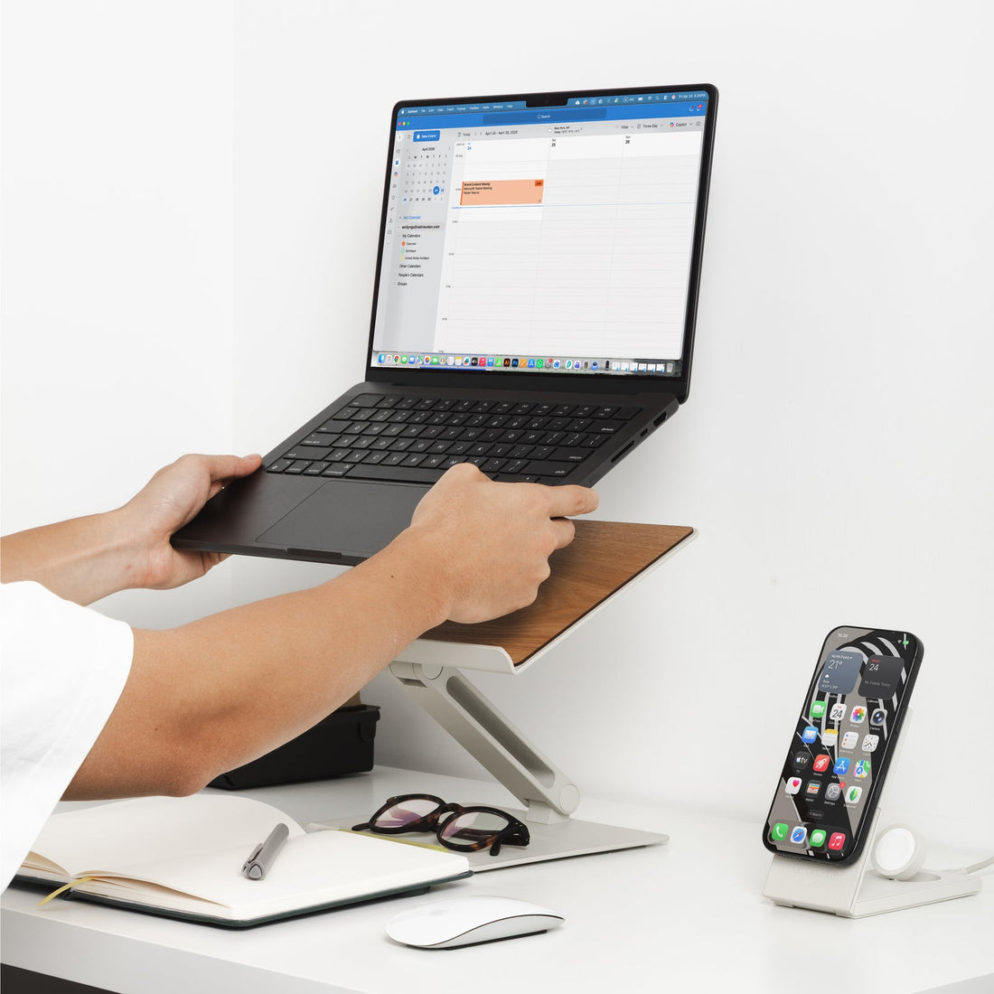 Person lifting a laptop onto the sandstone stand at a desk with a phone and notebook nearby