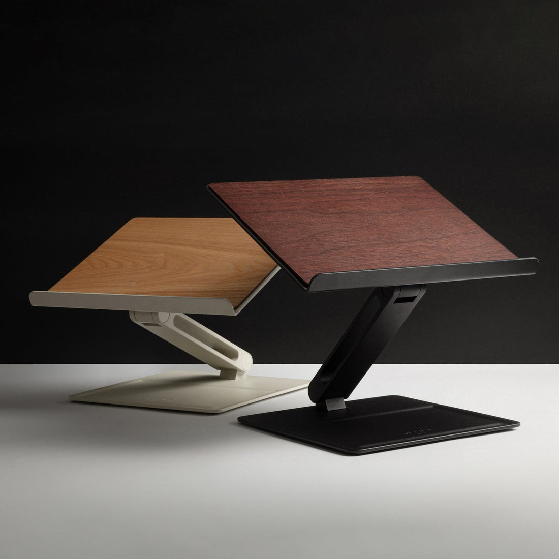 Two desk laptop stands, one with a light wood top and cream base and one with a dark wood top and black base, on a dark backdrop