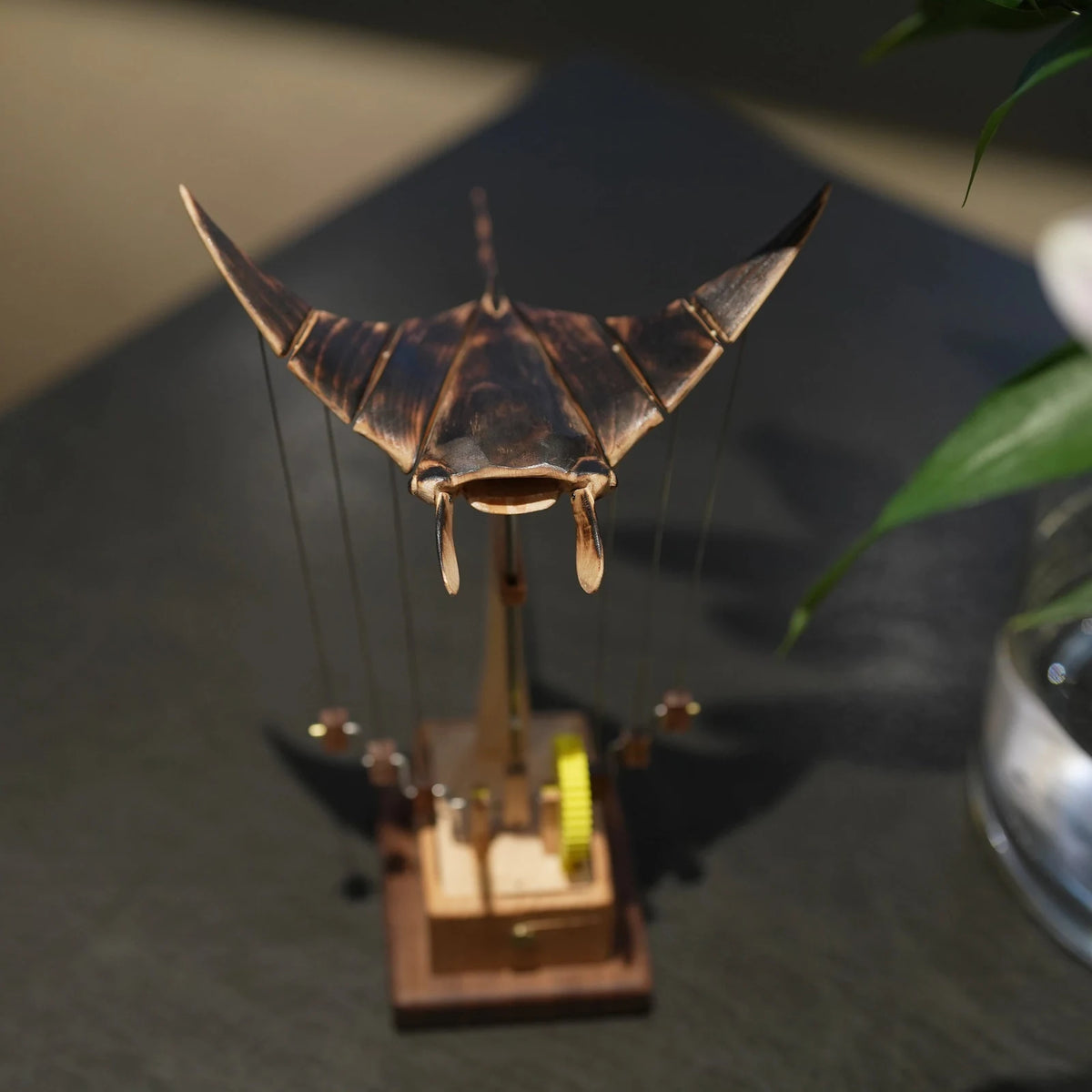 Dark ribbed manta ray automaton from the front above its tower, a green plant blurred on a dark table behind.