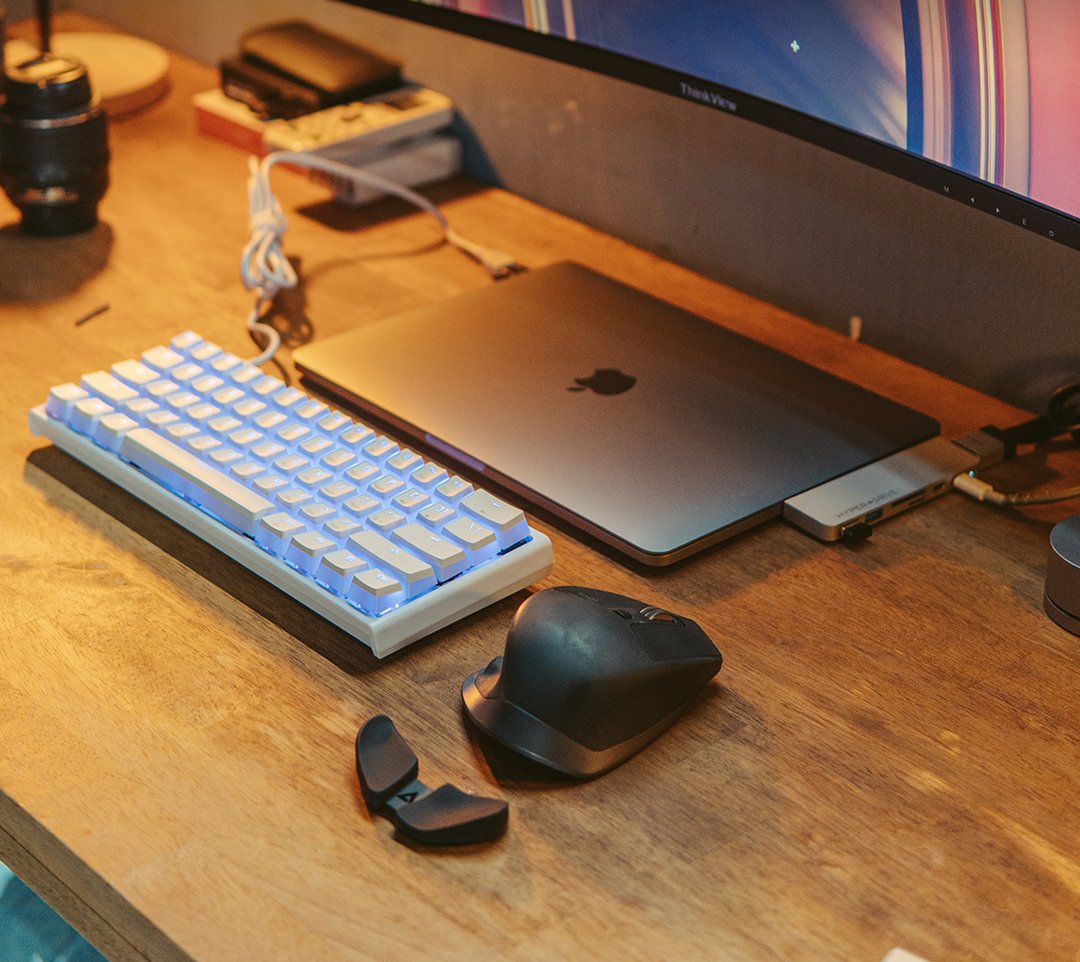 Wood desk with a white backlit keyboard, closed laptop and ergonomic mouse under a curved monitor