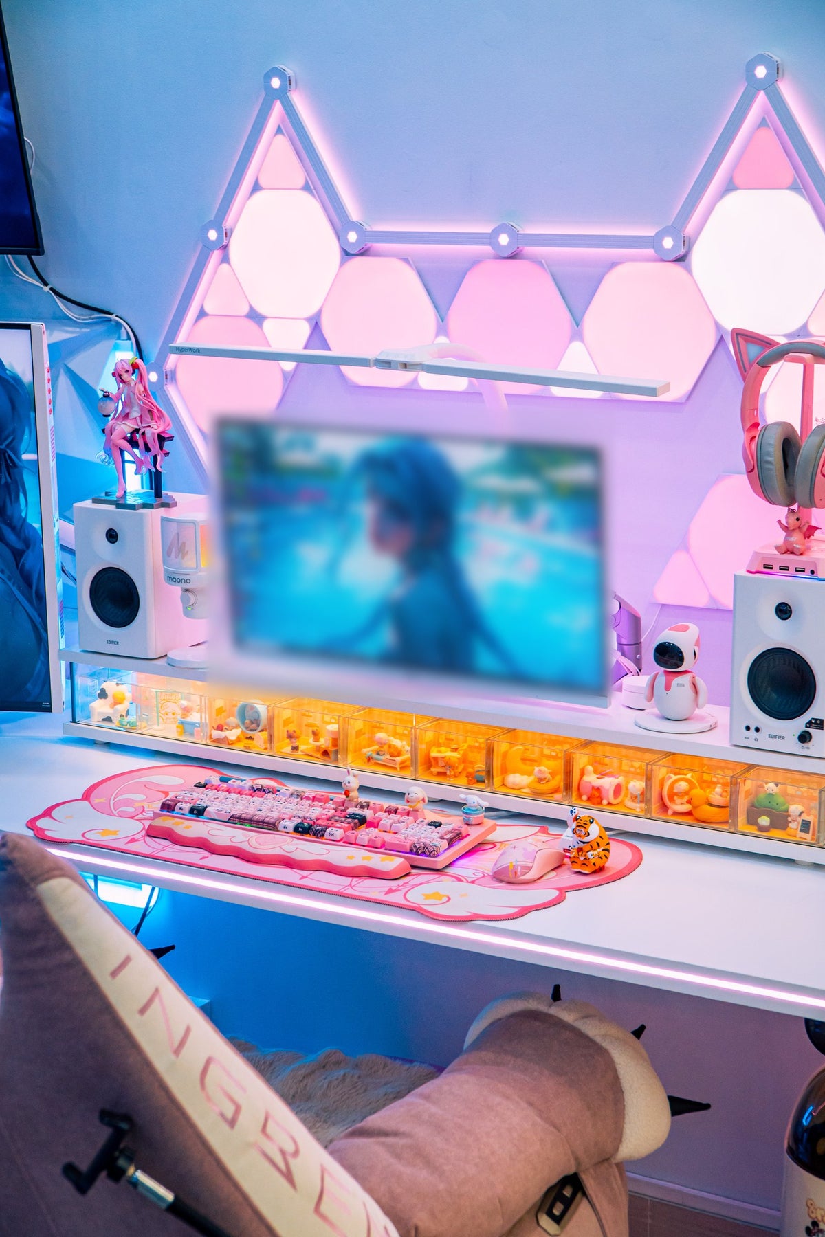 Front view of a white desk with a monitor, speakers, a light-up shelf of collectibles and a pink keyboard