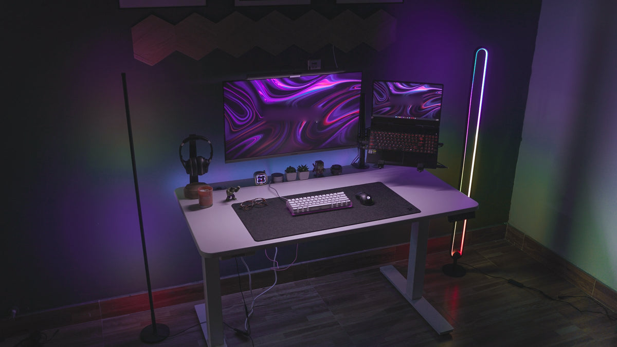 Dark purple-lit gaming desk with ultrawide monitor, laptop, headphone stand, keyboard, desk mat and neon floor lamps