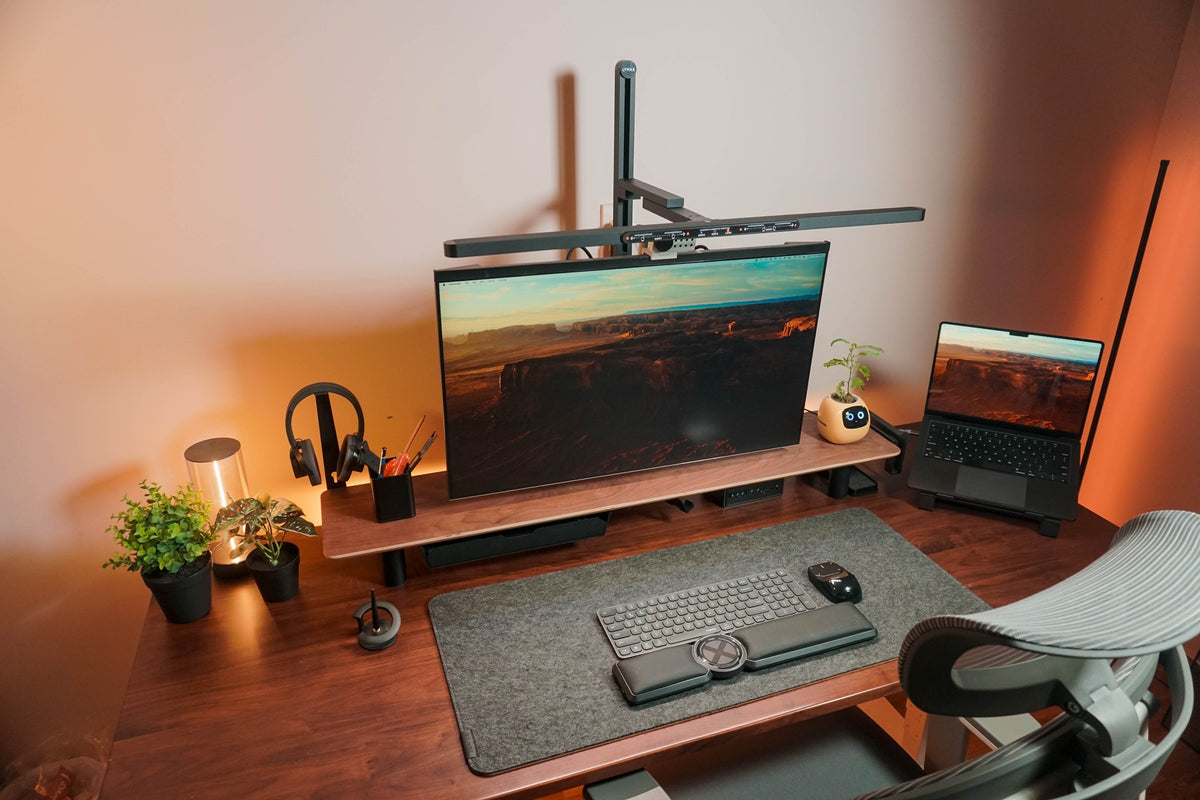 Walnut desk with a monitor shelf, felt mat, arm lamp and a laptop on a stand in warm evening light