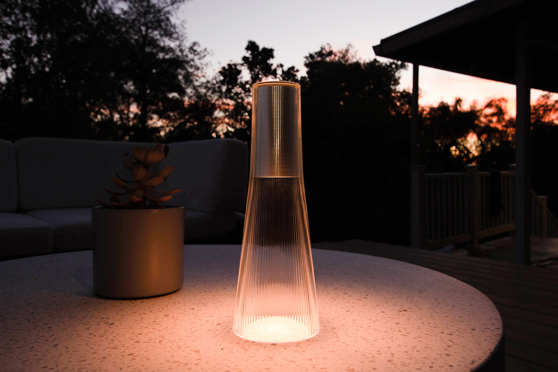 A bronze Candel lamp glowing on a round pale table at dusk with a plant behind