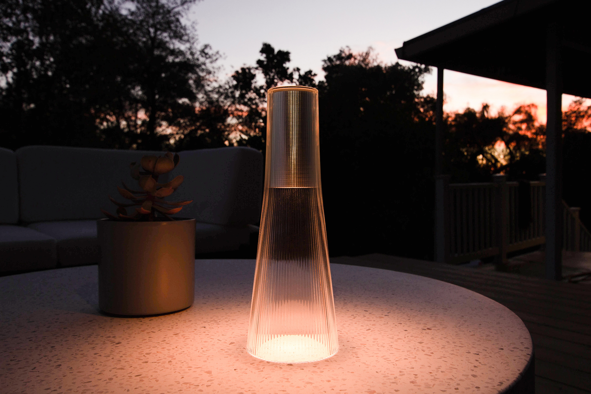 A bronze Candel lamp glowing on a round pale table at dusk with a plant behind