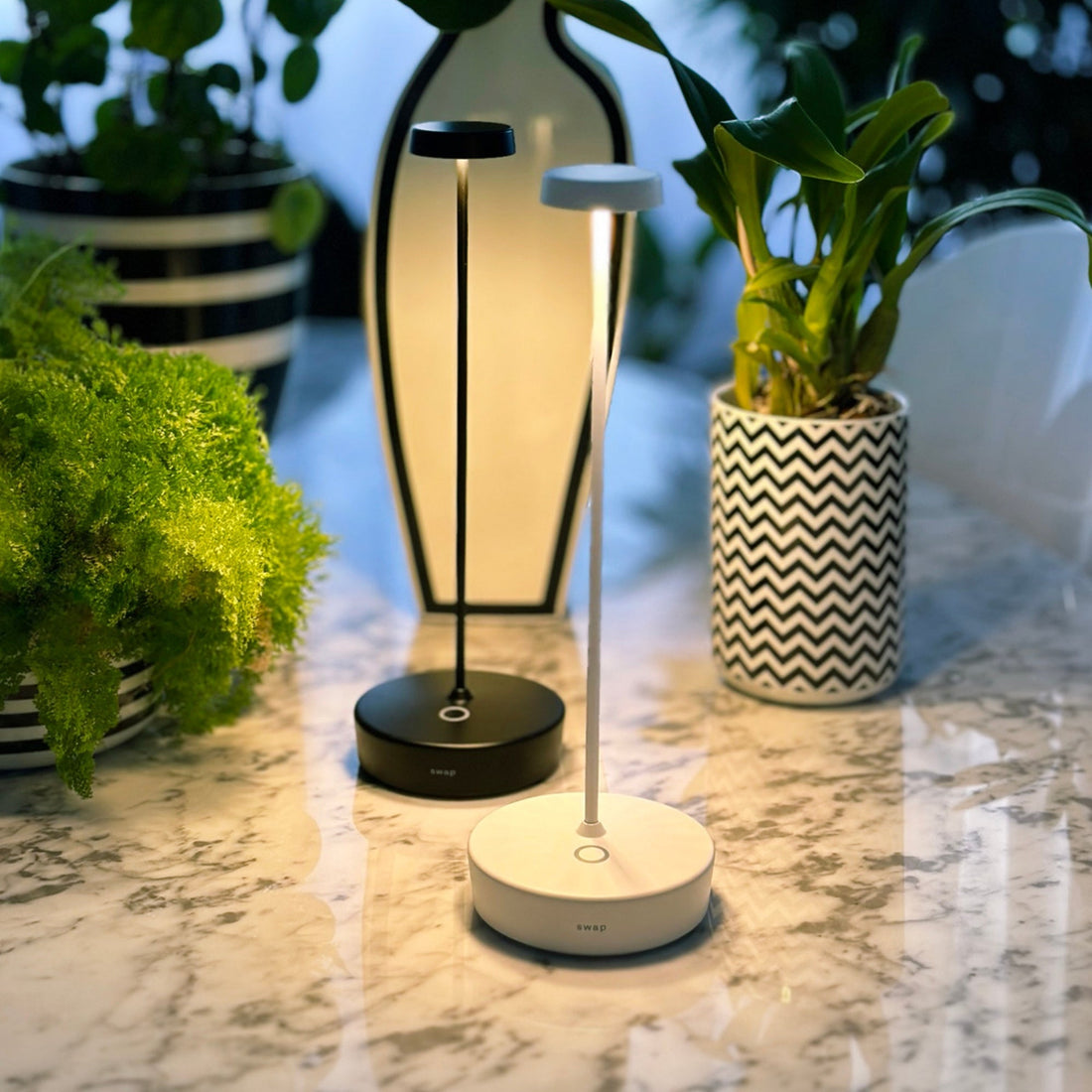 A black and a white Swap Pro lamp glowing on a marble surface beside patterned vases and plants
