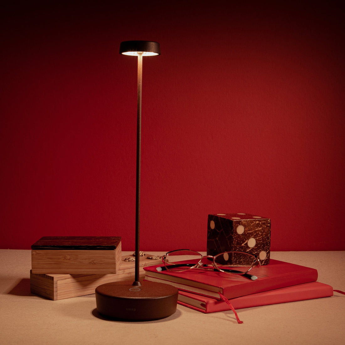 A black Swap Pro lamp lit against a red wall beside books and a pair of glasses