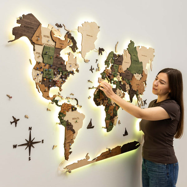 A person adjusting the wooden world map on a white wall with a warm yellow backlight glowing around the continents