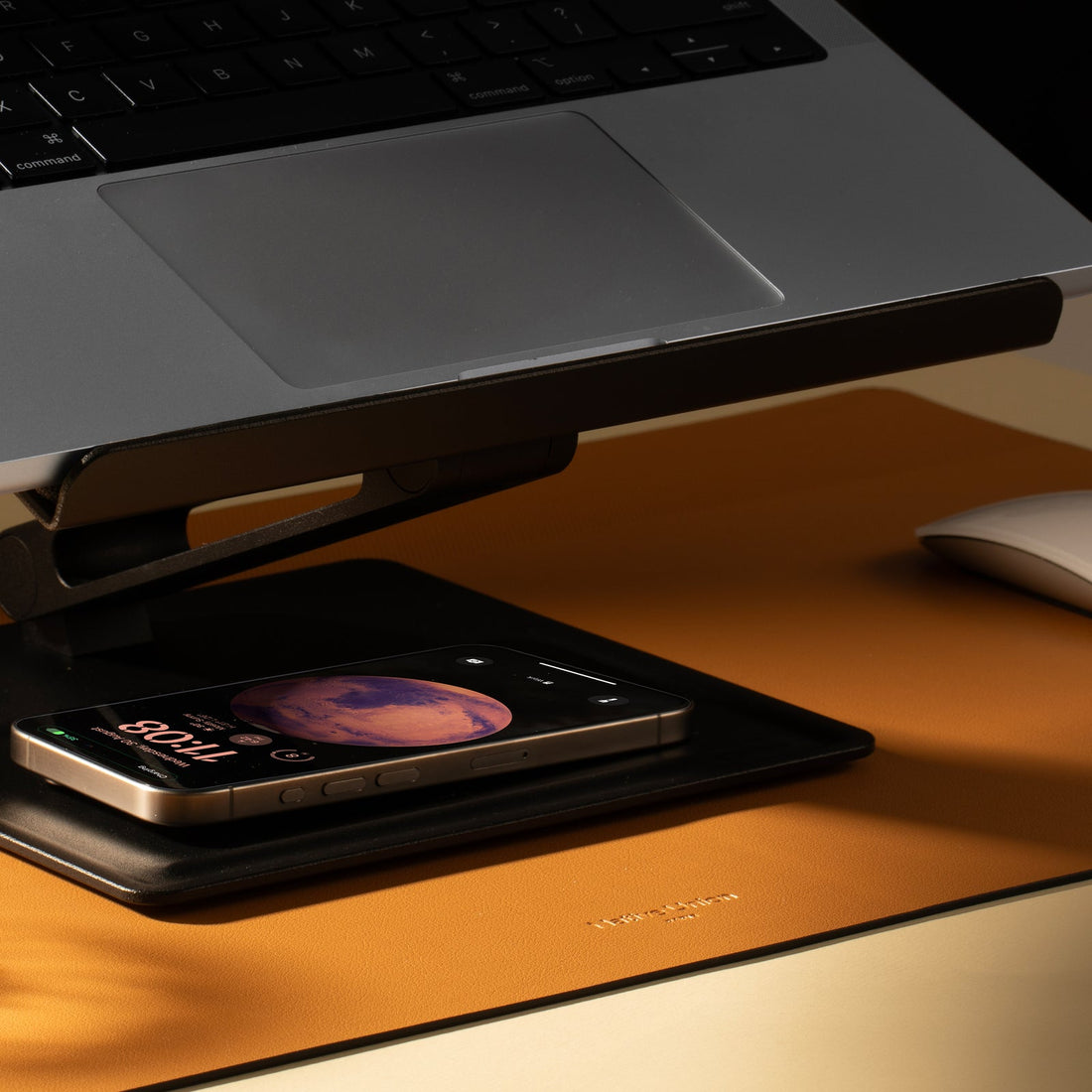 Laptop raised on the stand above a phone lying on the black base tray, on an orange desk mat