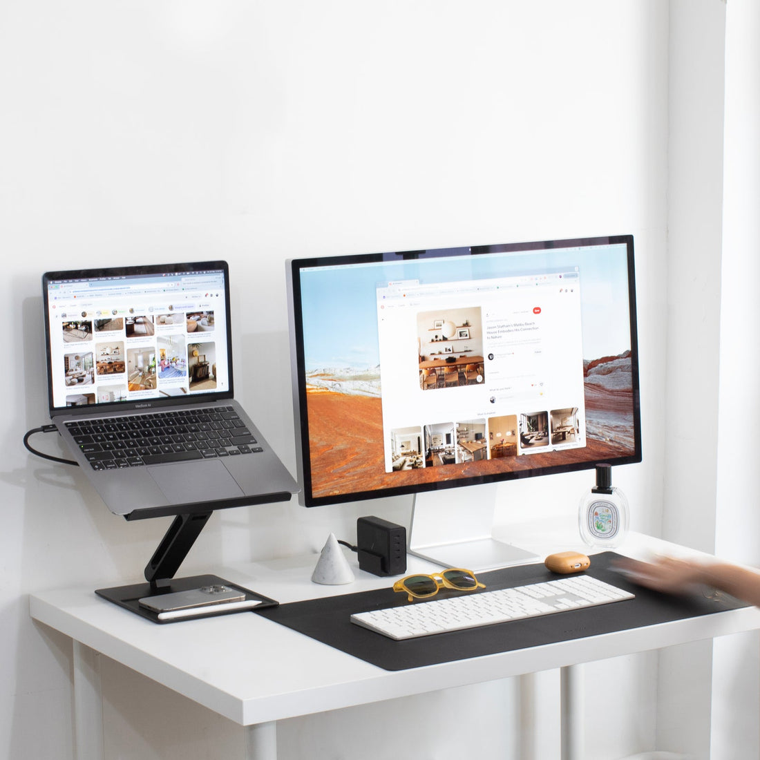 Desk with a laptop on the stand next to a monitor, on a white desk with a black desk mat