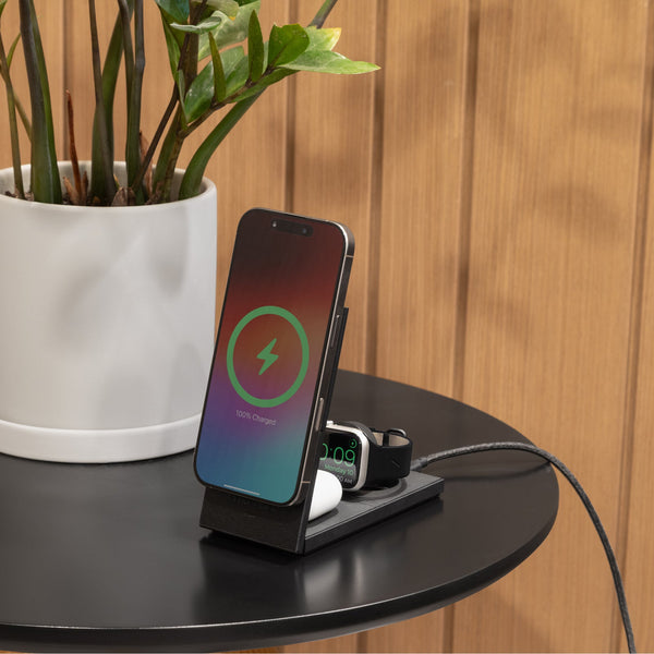 Black charging stand on a round table with a phone upright and a watch and earbuds on the base, a plant behind