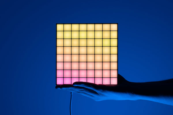 A hand holding one lit square LED panel with a warm yellow to pink gradient against a blue backdrop