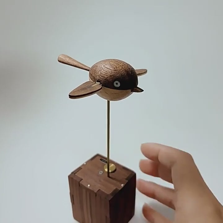 A hand reaches the brass button on top of the dark wood box and the round wooden bird on the brass rod above starts to beat its wings, plain light grey background.