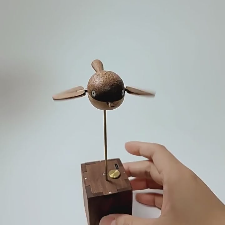 The wooden bird on its rod turning slowly while its wings beat, seen from the front and the side, the dark wood box with its brass button below.