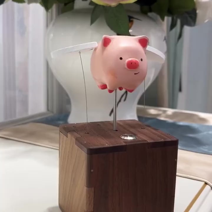 Pink pig with white wings on a slim rod above a dark wood box with a metal button on top, wings flapping up and down on thin wires, a white vase and a sheer curtain behind.