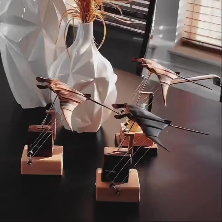 Three manta ray automata with different bases on a dark table beside a white vase, their wings moving.