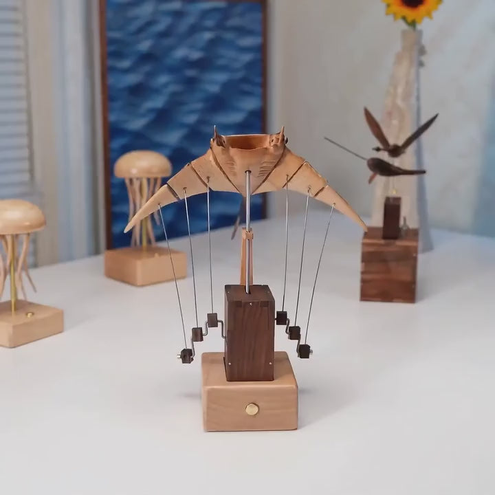Manta ray automaton with pale ribbed wings on thin wires above a dark wood tower and light wood base, a sunflower and small wooden jellyfish beside it on a white table.