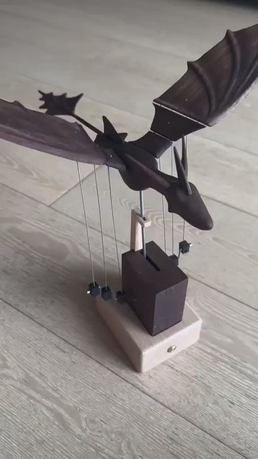 Dark dragon with wings spread on wires above a dark tower and pale base, standing on a light wood floor.