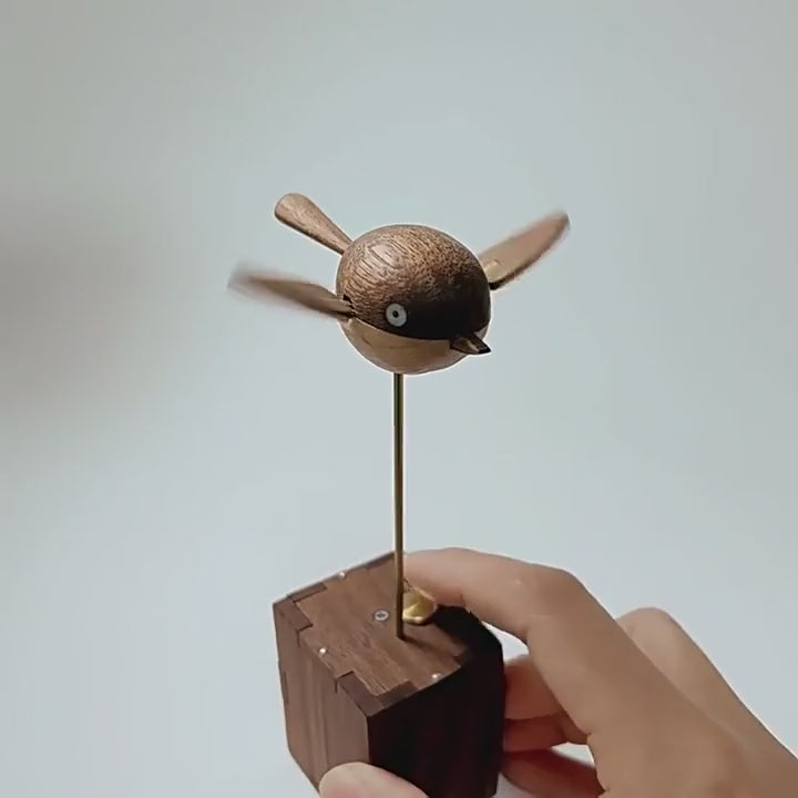 The round wooden bird beating both wings fast on the brass rod above the dark wood box, a hand resting beside the box, plain light grey background.