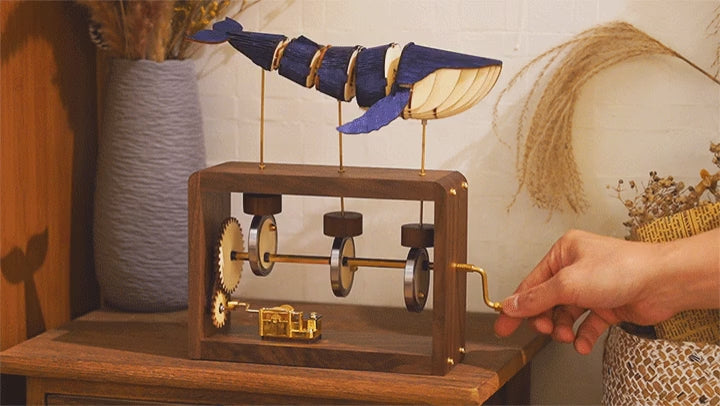 A hand turns the crank on the walnut edition and the blue-violet whale rolls on its rods, a vase of dried grass behind.