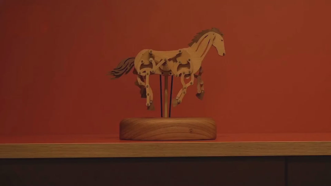 The jointed wooden horse galloping in place on its rods above a round wooden base against a red wall, from the whole piece to its moving legs and head.