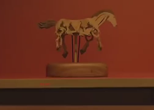 The jointed wooden horse galloping in place above its round base on a dark shelf, red wall behind.