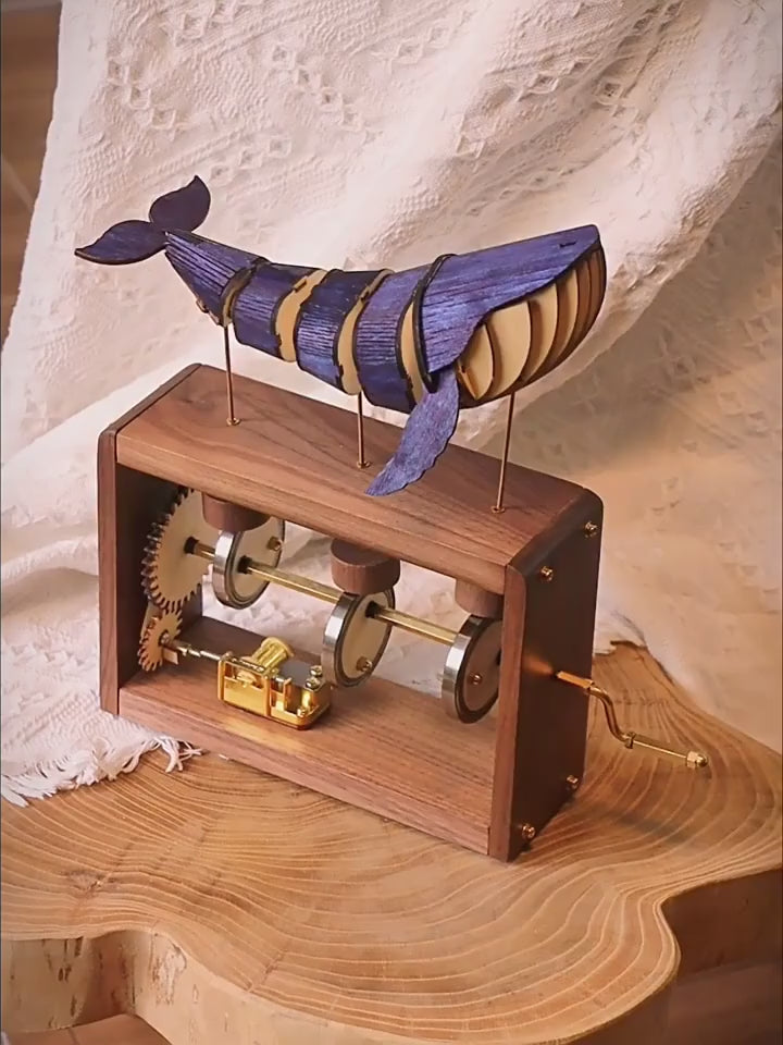 A hand turns the brass crank on a dark wood music box; a blue-violet jointed wooden whale rolls on three brass rods above, gears and cams turning inside the open frame.