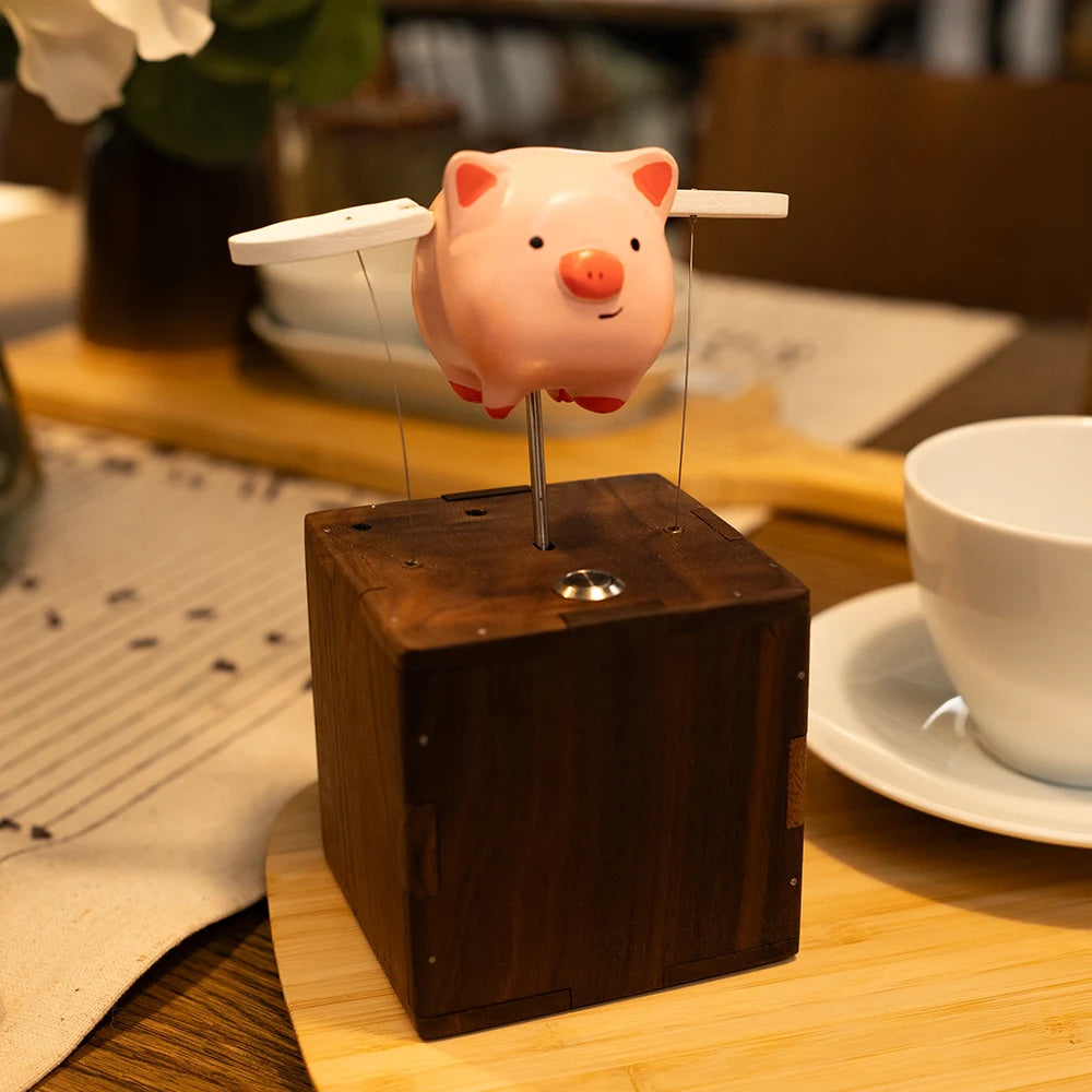 Pink pig with white wings on a slim rod above a dark wood box, set on a bamboo board beside a white cup and saucer.