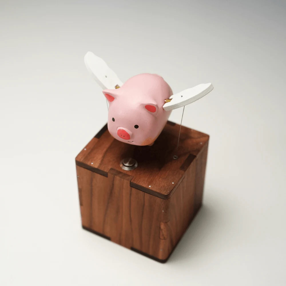 Pink pig tipped to one side with white wings spread, held on a slim rod above a reddish-brown wood box with finger-jointed corners and a metal button, plain pale background.