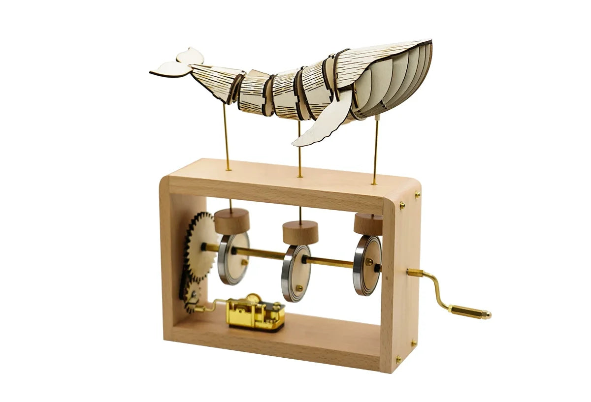 Pale wood frame music box on white: a pale jointed whale on three brass rods, gear wheel, three cams, gold music box movement and a brass crank on the right.