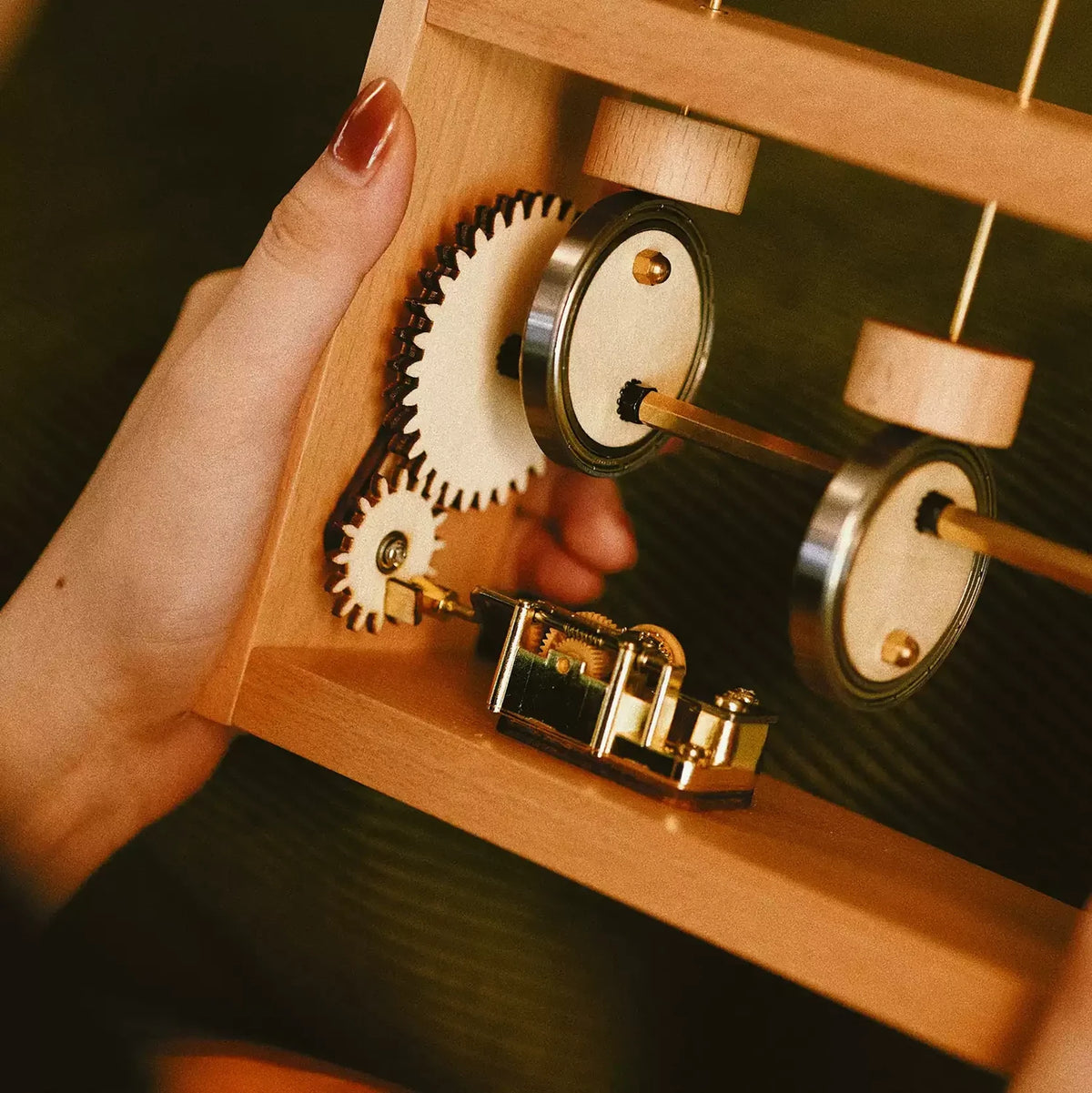Close view of a pale wood frame held in one hand: the wooden gear wheels, cams on a brass shaft and the gold music box movement.