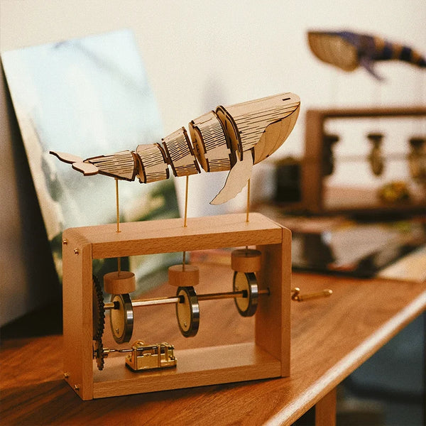 Pale wood music box on a wooden shelf: a pale jointed wooden whale on three brass rods above the open frame, its gear wheel, three cams and the gold music box movement in view.
