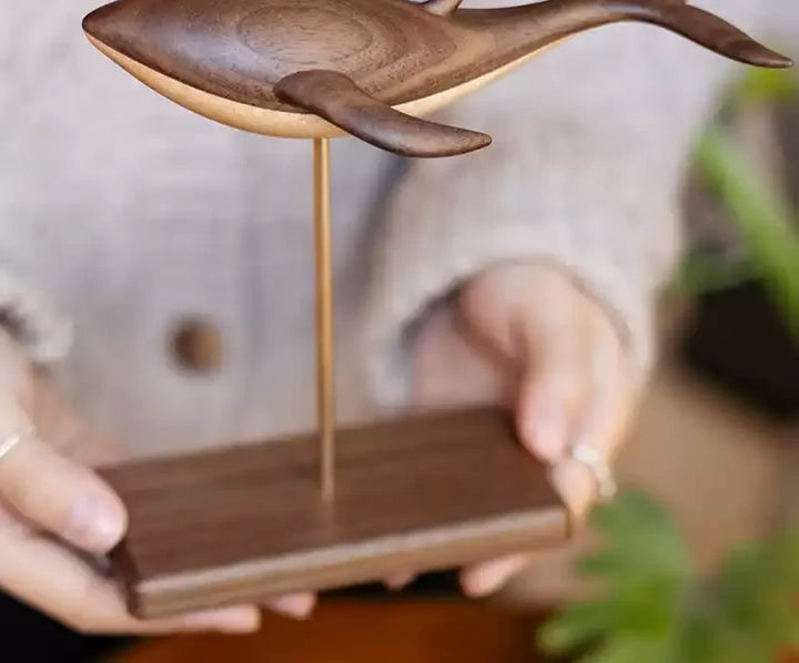 Two hands hold the standing edition: a walnut whale on a slim brass rod above a dark walnut stand.