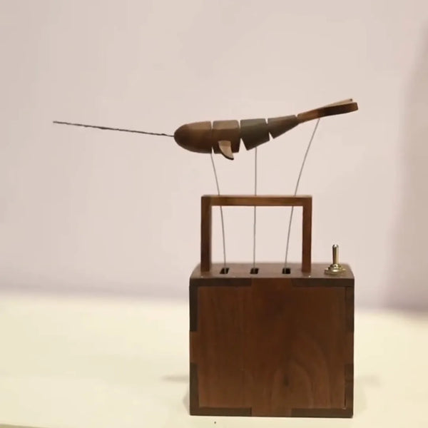 Front view of the dark wood whale hanging from three wires above an open dark wood frame on a finger-jointed dark wood box, small toggle switch at the right of the lid, pale wall behind.