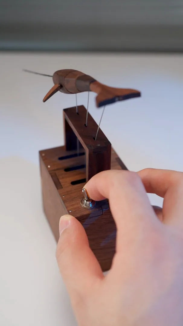 A finger flipping the small metal toggle switch on the dark wood box, whale hanging on wires above the open dark wood frame.